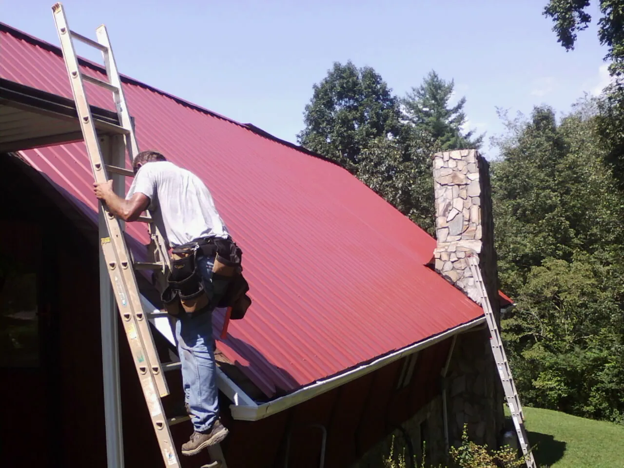 Roofer on a ladder beside a red metal roof and stone chimney — Soffit & Fascia Repair by Roofing Brothers in Childersburg